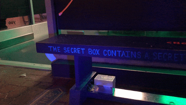 A scrolling LED sign on the exterior bench reading The Secret Box Contains A Secret